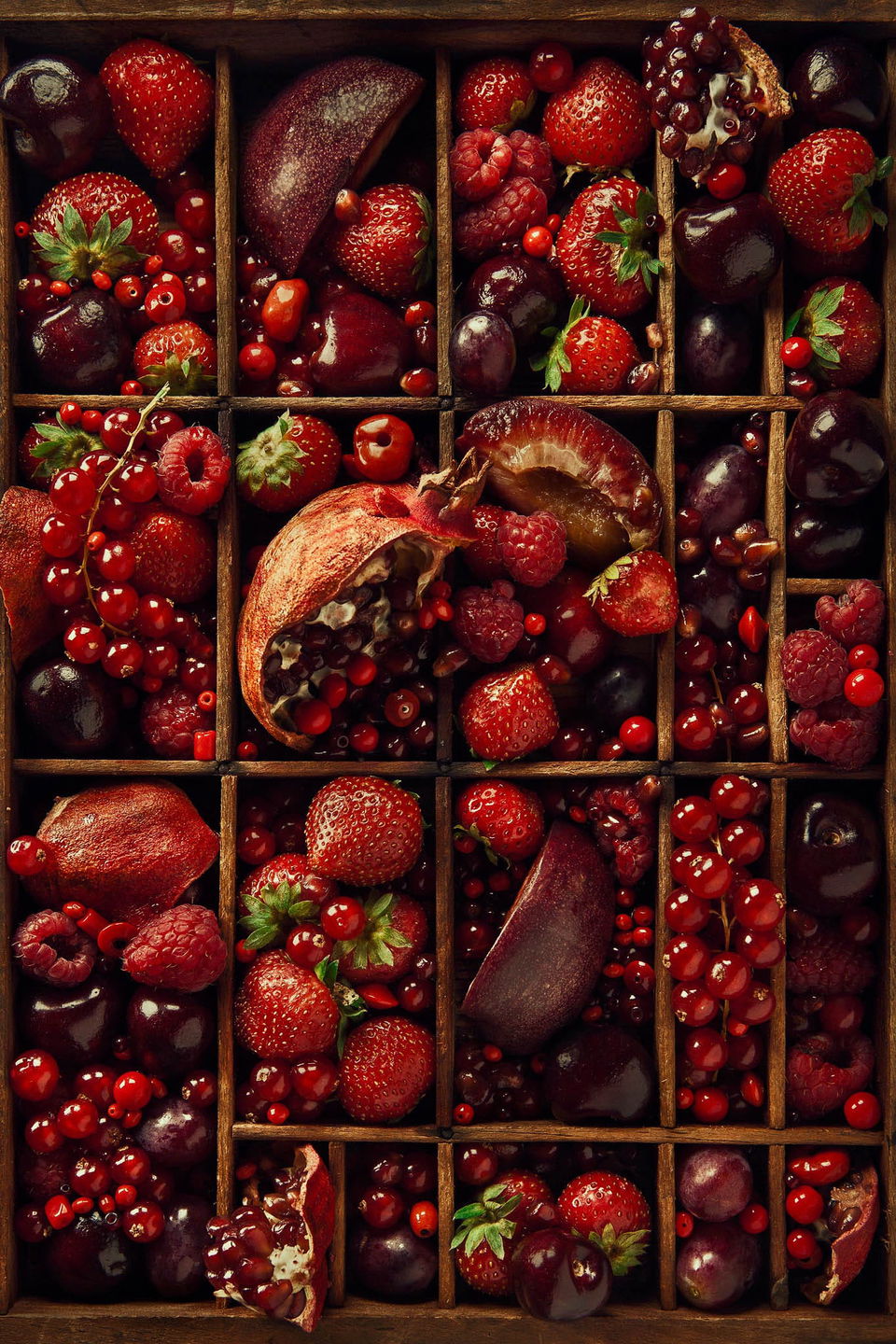 Berries and pomegranate arranged in wooden compartments, glossy dark-red tones and dramatic light